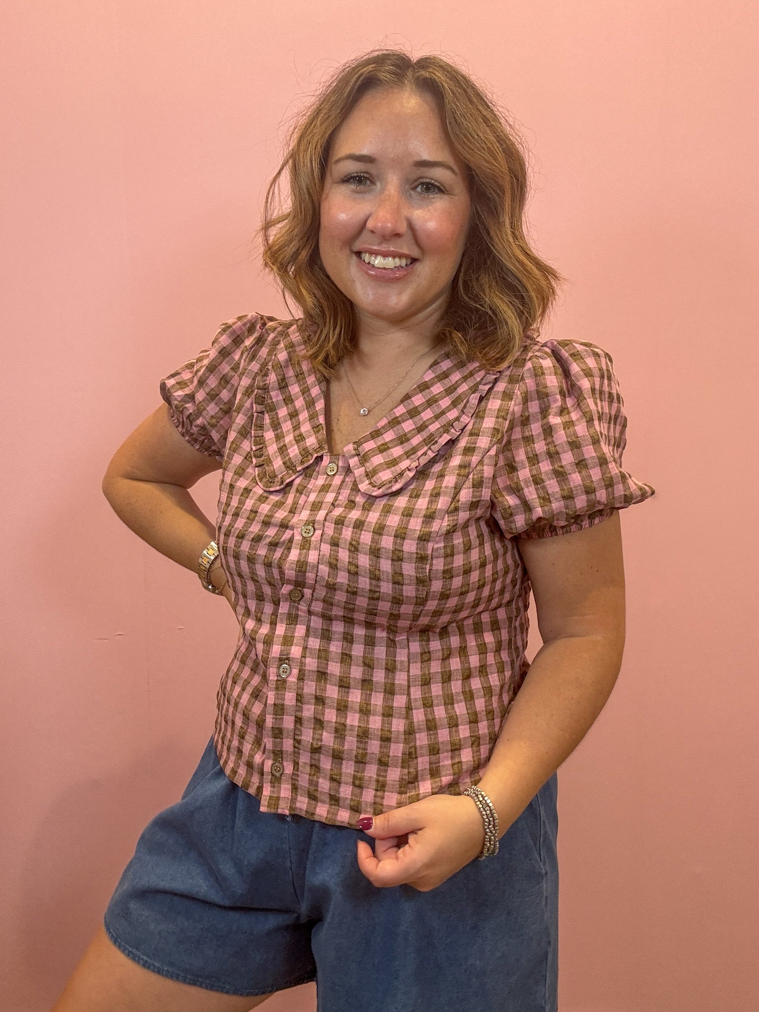 A woman wearing a pink and brown gingham short-sleeve button-up shirt with puff sleeves and a Peter Pan collar, paired with blue denim shorts, posing against a solid pink wall.