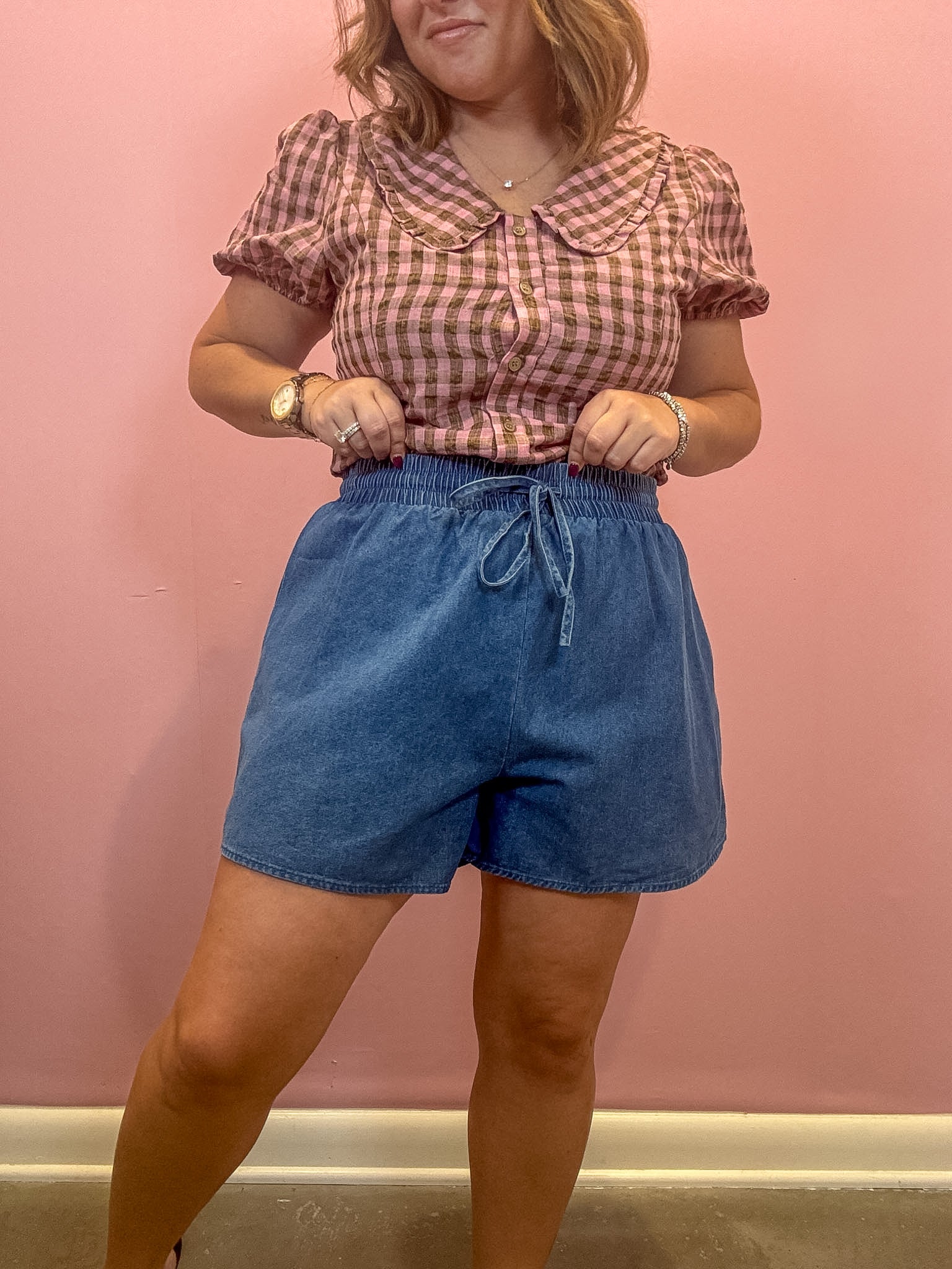 A woman modeling a pink and brown gingham short-sleeve blouse with a Peter Pan collar paired with blue denim drawstring shorts, standing against a solid pink wall.