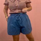 A woman modeling a pink and white gingham short-sleeve blouse with a Peter Pan collar and blue denim shorts, standing against a solid pink wall.