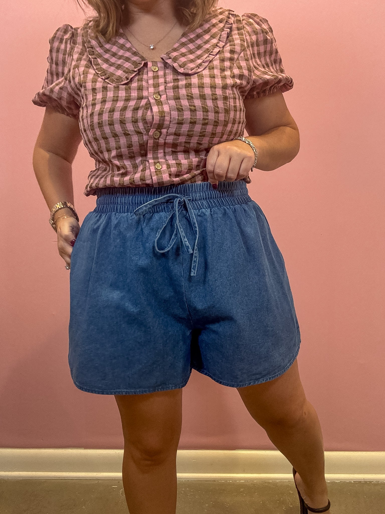 A woman modeling a pink and white gingham short-sleeve blouse with a Peter Pan collar and blue denim shorts, standing against a solid pink wall.