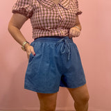 A woman modeling a pink and white gingham short-sleeve blouse with a Peter Pan collar paired with blue denim drawstring shorts, standing against a solid pink wall.