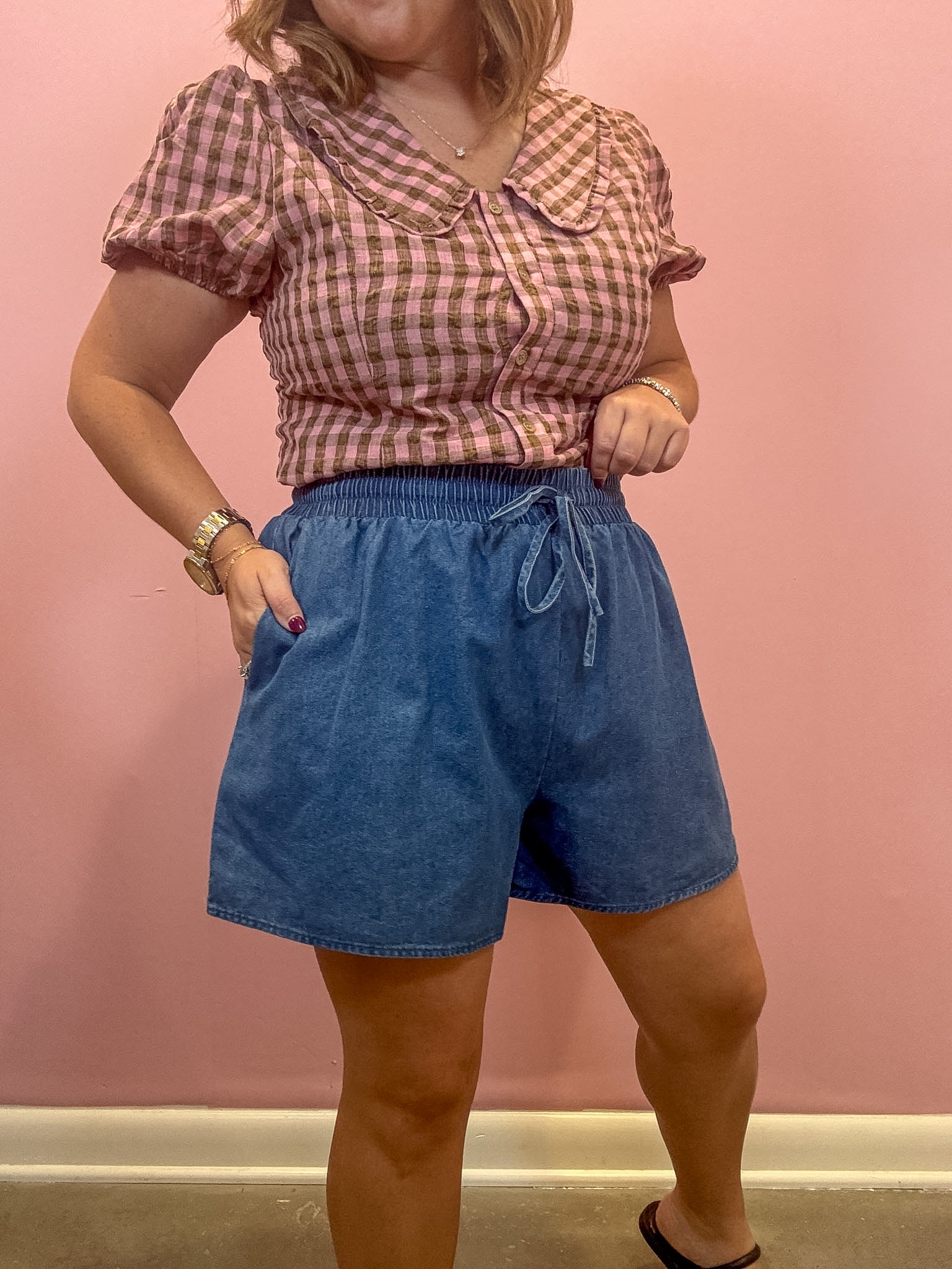A woman modeling a pink and white gingham short-sleeve blouse with a Peter Pan collar paired with blue denim drawstring shorts, standing against a solid pink wall.