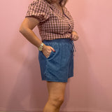 A woman modeling a pink and brown gingham short-sleeve top and blue denim shorts against a pink wall.