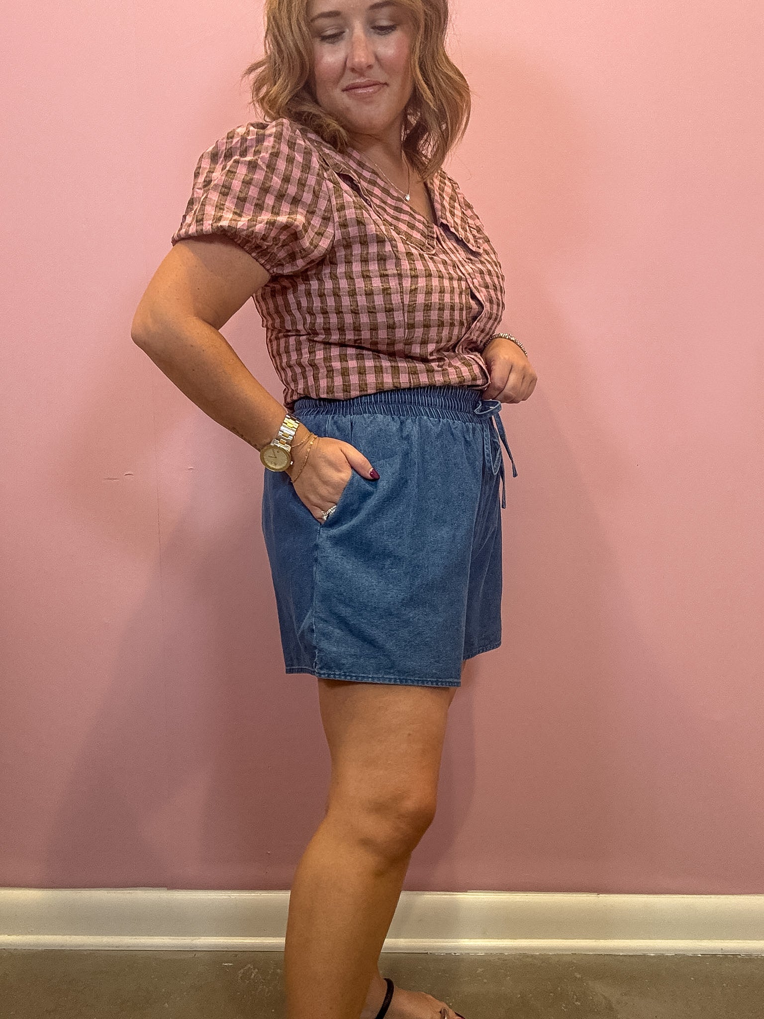 A woman modeling a pink and brown gingham short-sleeve top and blue denim shorts against a pink wall.