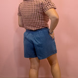 A woman shown from the back wearing a pink and brown gingham short-sleeve shirt and blue denim shorts, standing against a solid pink wall.