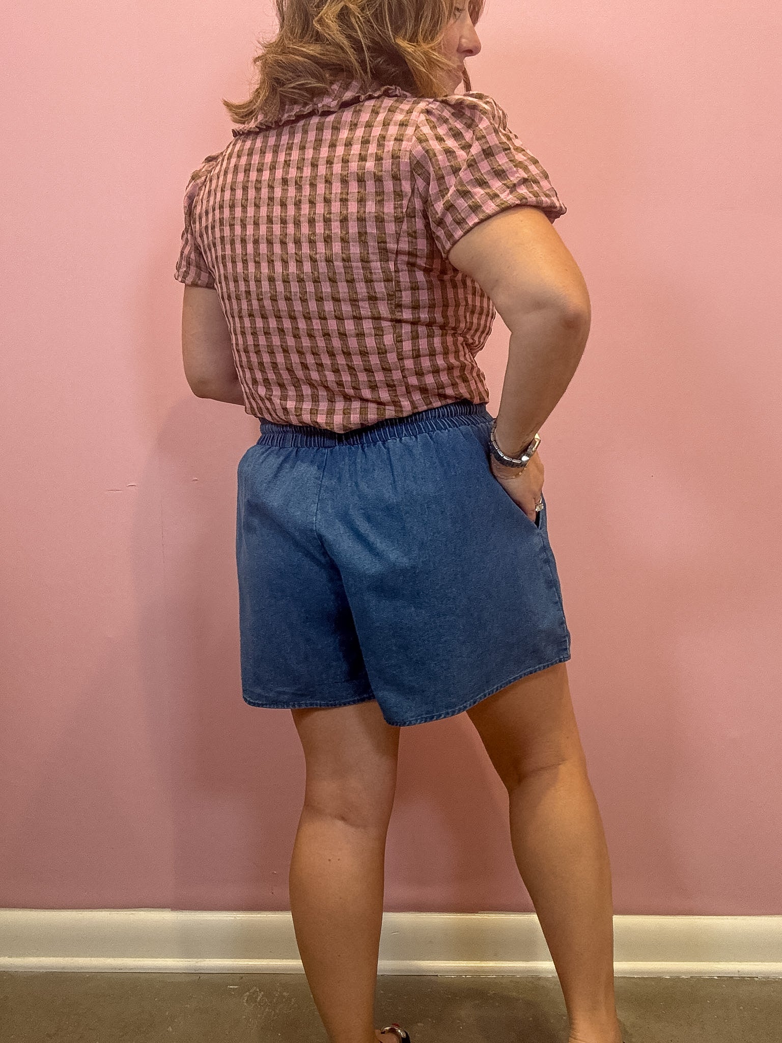 A woman shown from the back wearing a pink and brown gingham short-sleeve shirt and blue denim shorts, standing against a solid pink wall.