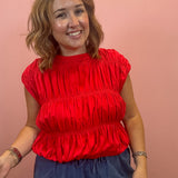 A woman with shoulder-length brown hair smiling, wearing a bright red ruched short-sleeve top and dark blue drawstring pants, standing against a solid pink wall.