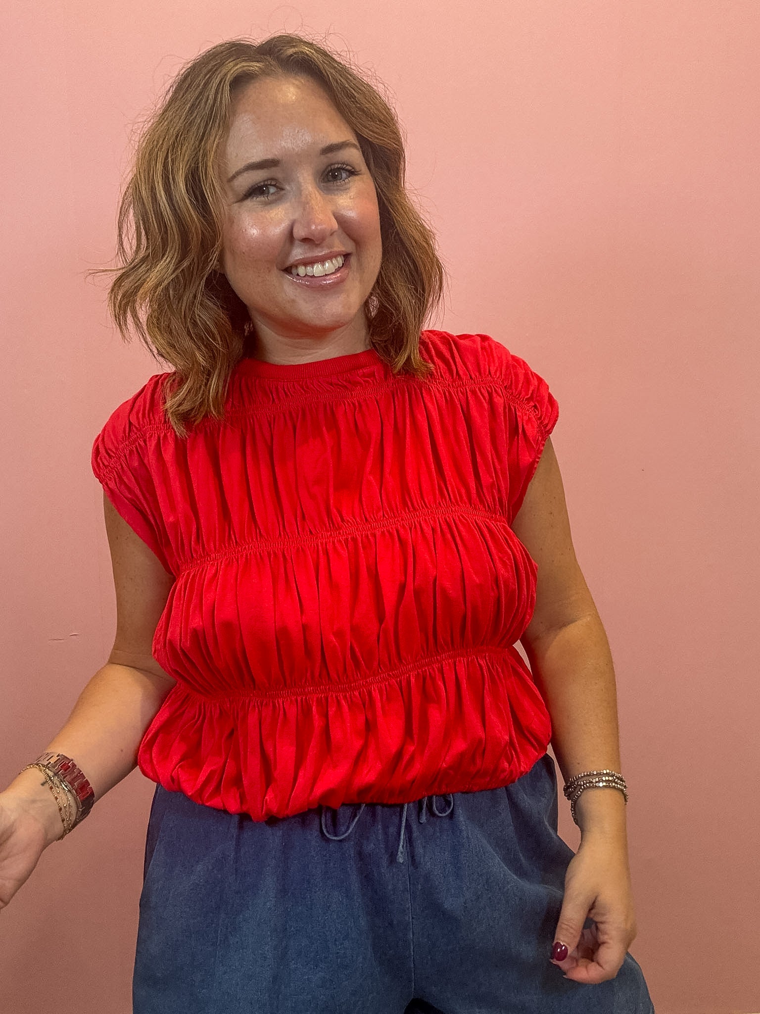 A woman with shoulder-length brown hair smiling, wearing a bright red ruched short-sleeve top and dark blue drawstring pants, standing against a solid pink wall.