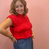 A woman wearing a bright red sleeveless smocked blouse and a blue denim skirt, posing against a solid pink background.