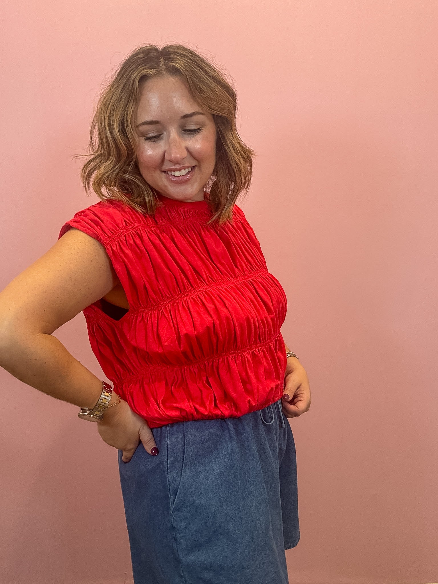 A woman wearing a bright red sleeveless smocked blouse and a blue denim skirt, posing against a solid pink background.