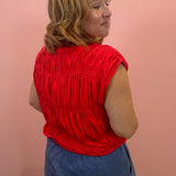 A woman wearing a bright red sleeveless top with horizontal ruching and blue denim shorts, standing against a solid pink wall.