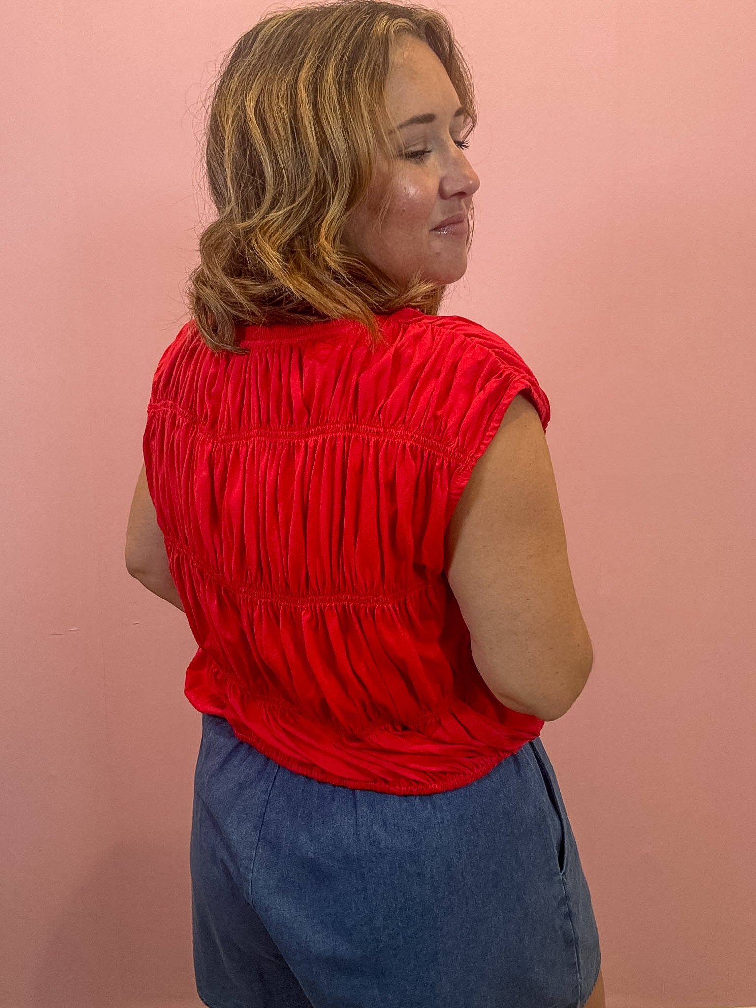 A woman wearing a bright red sleeveless top with horizontal ruching and blue denim shorts, standing against a solid pink wall.