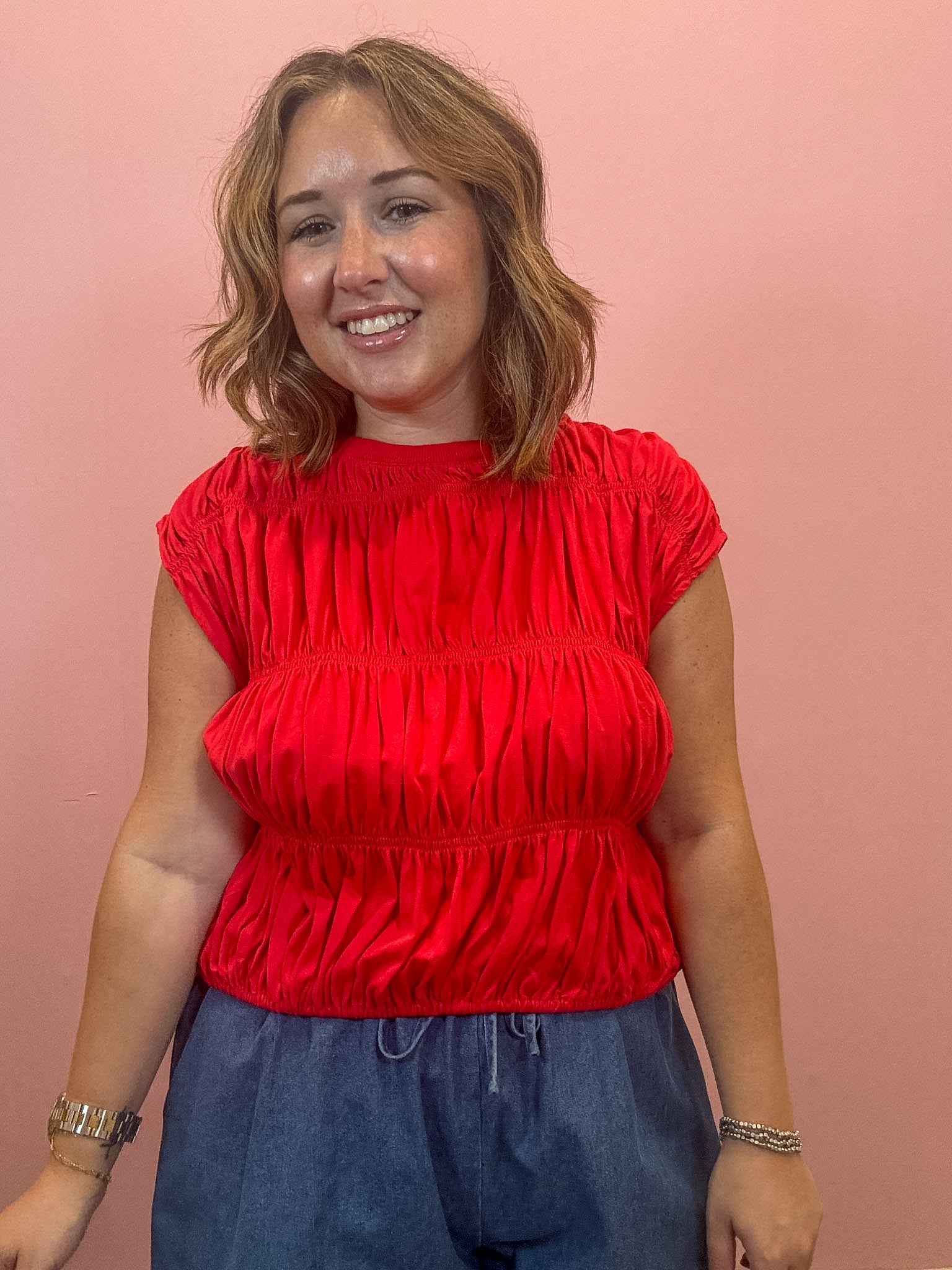 A woman with shoulder-length light brown hair smiling, wearing a bright red ruched short-sleeve top and blue drawstring pants, standing against a solid pink wall.