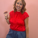 A smiling woman with shoulder-length brown hair stands against a solid pink wall, wearing a bright red ruched short-sleeve top and blue denim shorts.