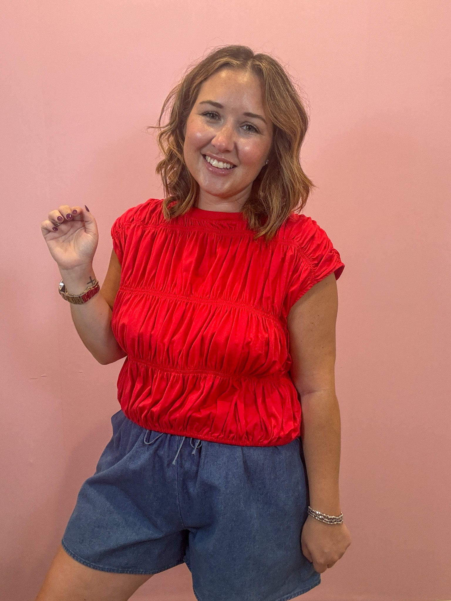A smiling woman with shoulder-length brown hair stands against a solid pink wall, wearing a bright red ruched short-sleeve top and blue denim shorts.