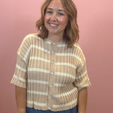 A woman with shoulder-length brown hair smiling, wearing a beige and white striped short-sleeve ribbed knit top with buttons, paired with blue jeans, standing against a solid pink wall.