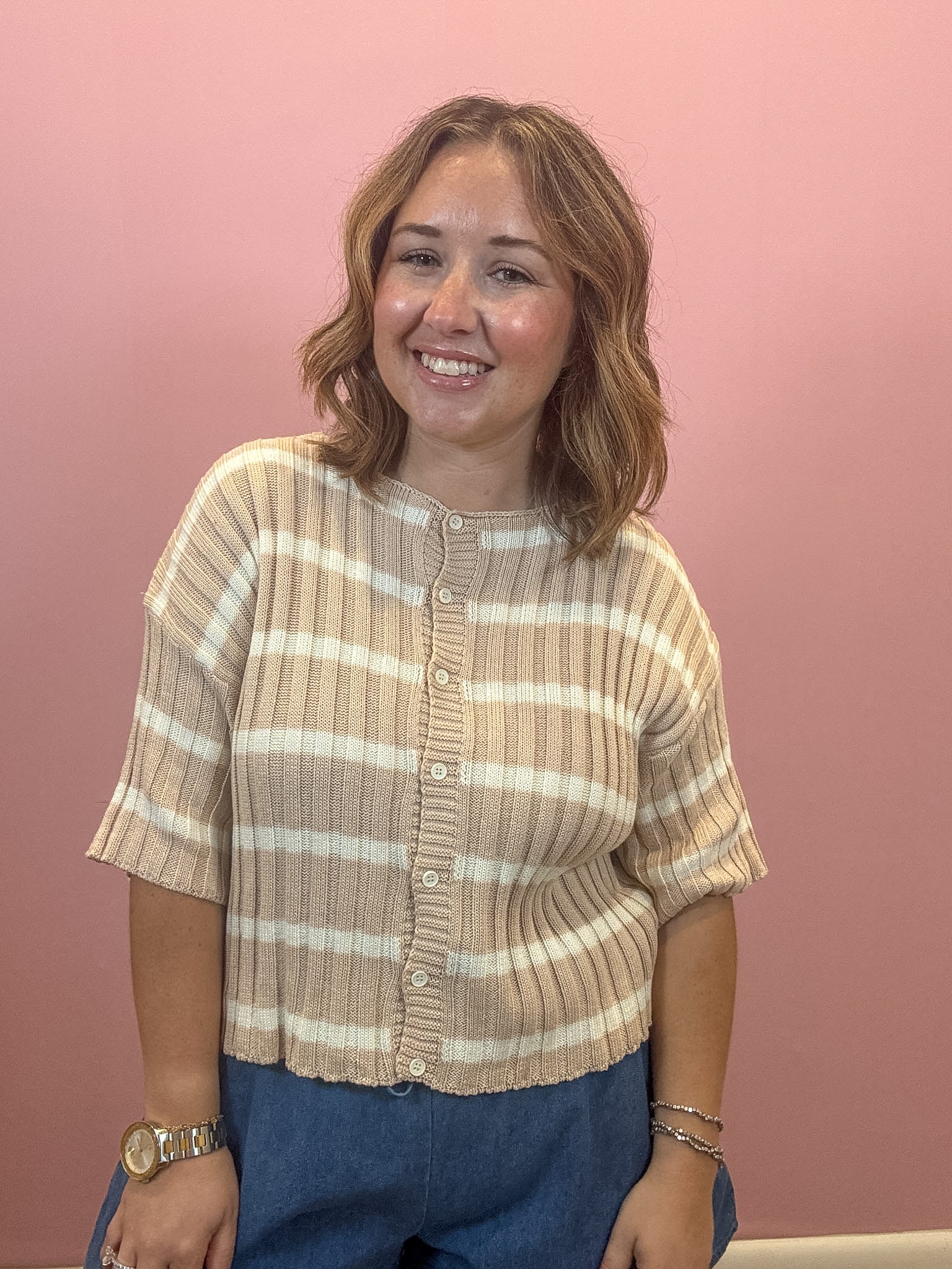 A woman with shoulder-length brown hair smiling, wearing a beige and white striped short-sleeve ribbed knit top with buttons, paired with blue jeans, standing against a solid pink wall.