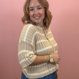 A woman with shoulder-length brown hair smiling and posing in a beige and white striped ribbed knit top, paired with blue jeans, against a solid pink wall.