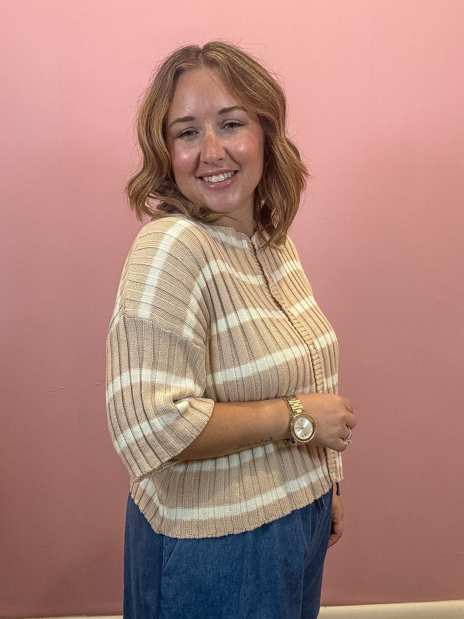 A woman with shoulder-length brown hair smiling and posing in a beige and white striped ribbed knit top, paired with blue jeans, against a solid pink wall.