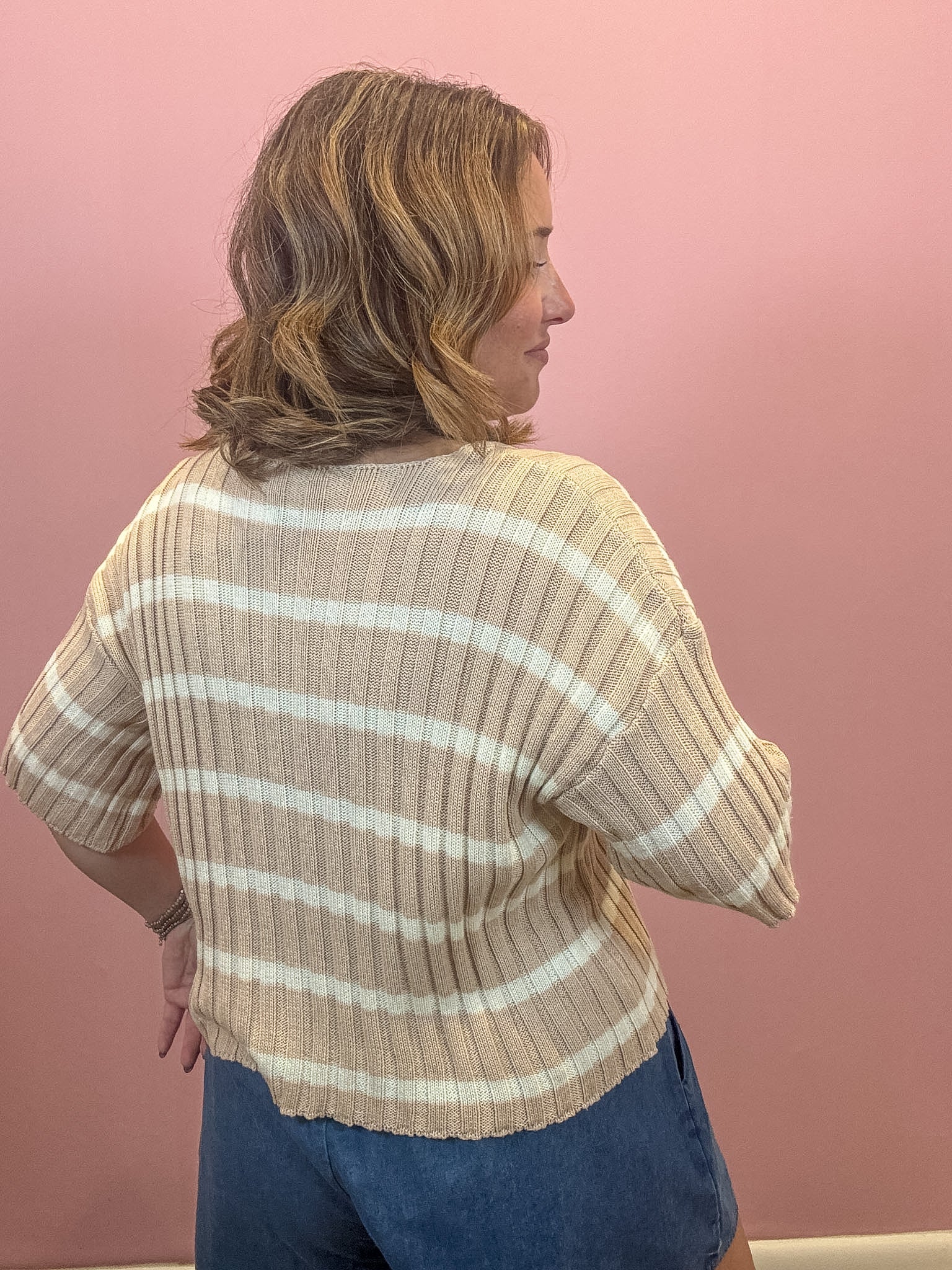 A woman shown from the back wearing a beige and white horizontally striped ribbed knit top, paired with blue jeans, against a solid pink wall.
