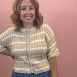 A smiling woman with shoulder-length wavy brown hair stands against a solid dusty pink wall, wearing a beige and white horizontally striped ribbed knit cardigan with short sleeves and blue pants.