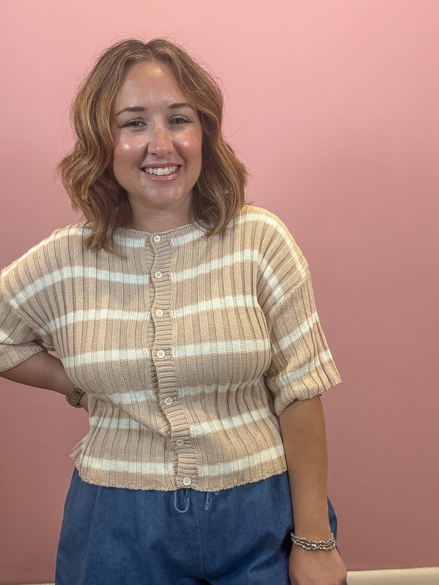 A smiling woman with shoulder-length wavy brown hair stands against a solid dusty pink wall, wearing a beige and white horizontally striped ribbed knit cardigan with short sleeves and blue pants.