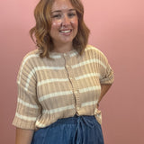 A woman with shoulder-length wavy brown hair smiling, wearing a beige and cream horizontally striped short-sleeve ribbed knit top with a button placket, paired with blue denim drawstring pants, set against a solid dusty pink wall.