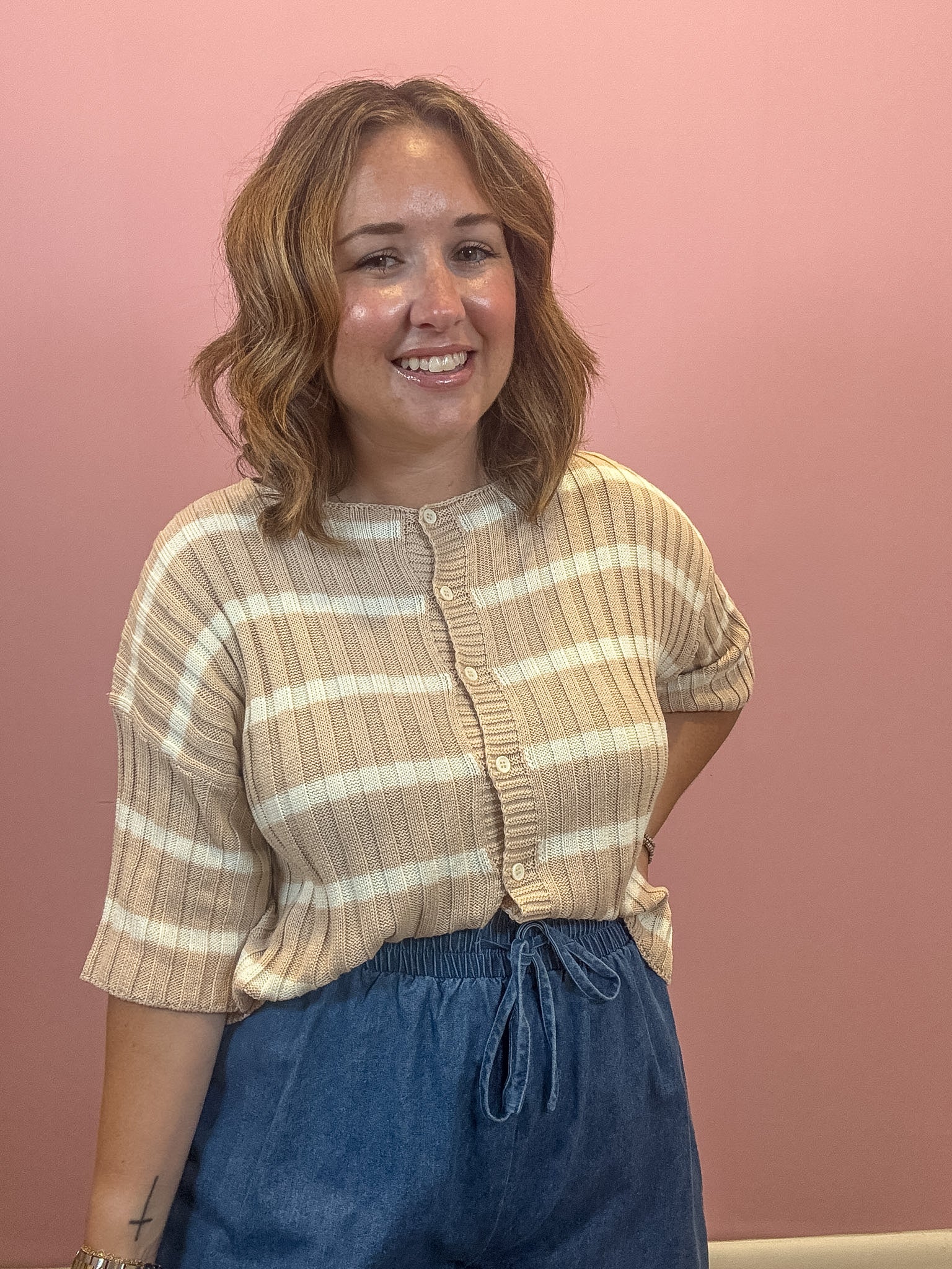 A woman with shoulder-length wavy brown hair smiling, wearing a beige and cream horizontally striped short-sleeve ribbed knit top with a button placket, paired with blue denim drawstring pants, set against a solid dusty pink wall.