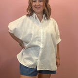 A woman modeling a white textured short-sleeve button-up shirt with blue denim shorts, standing against a solid pink wall.
