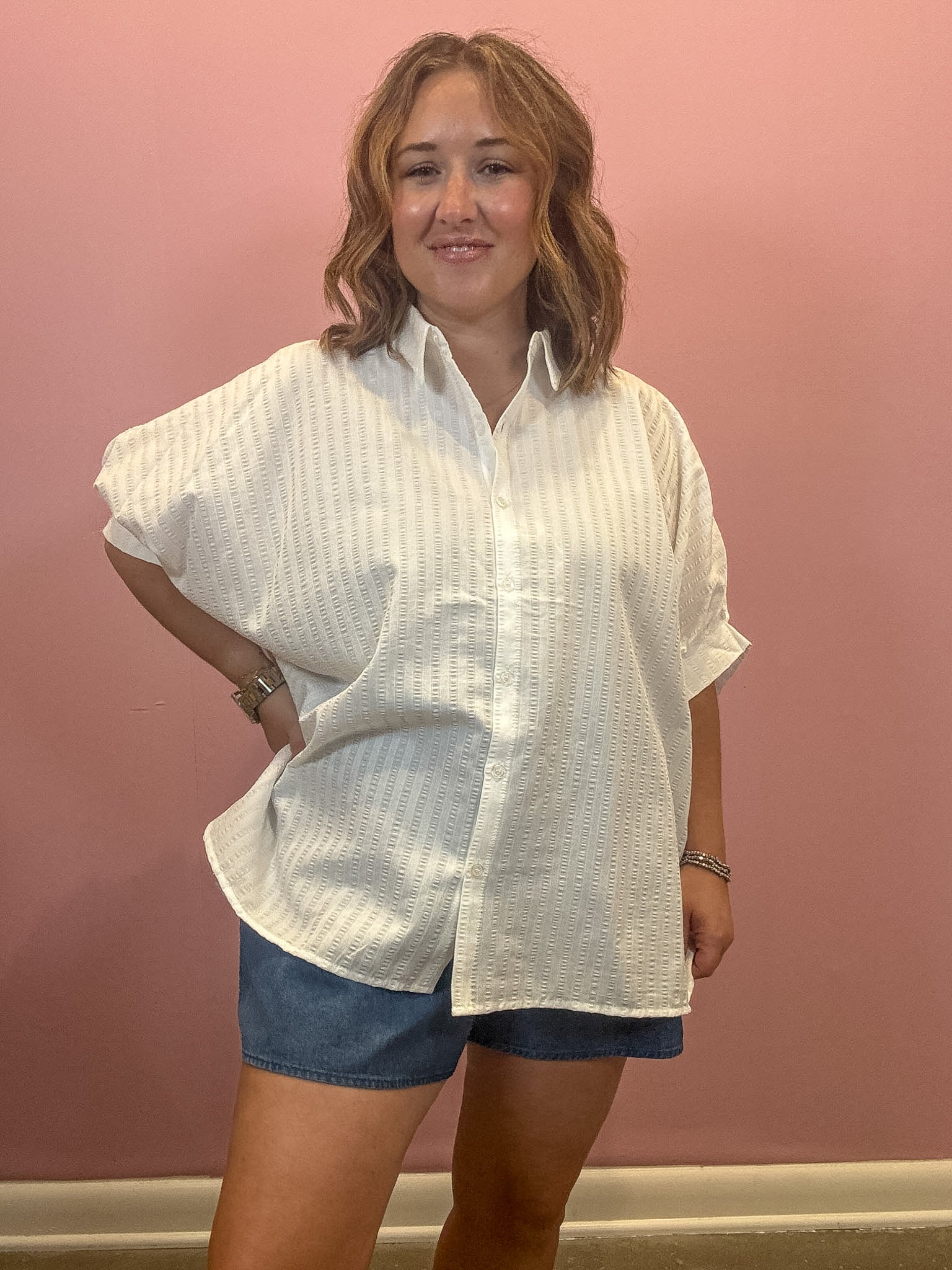 A woman modeling a white textured short-sleeve button-up shirt with blue denim shorts, standing against a solid pink wall.