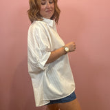 A woman wearing an oversized white textured short-sleeve button-up shirt and denim shorts, standing against a solid pink wall.