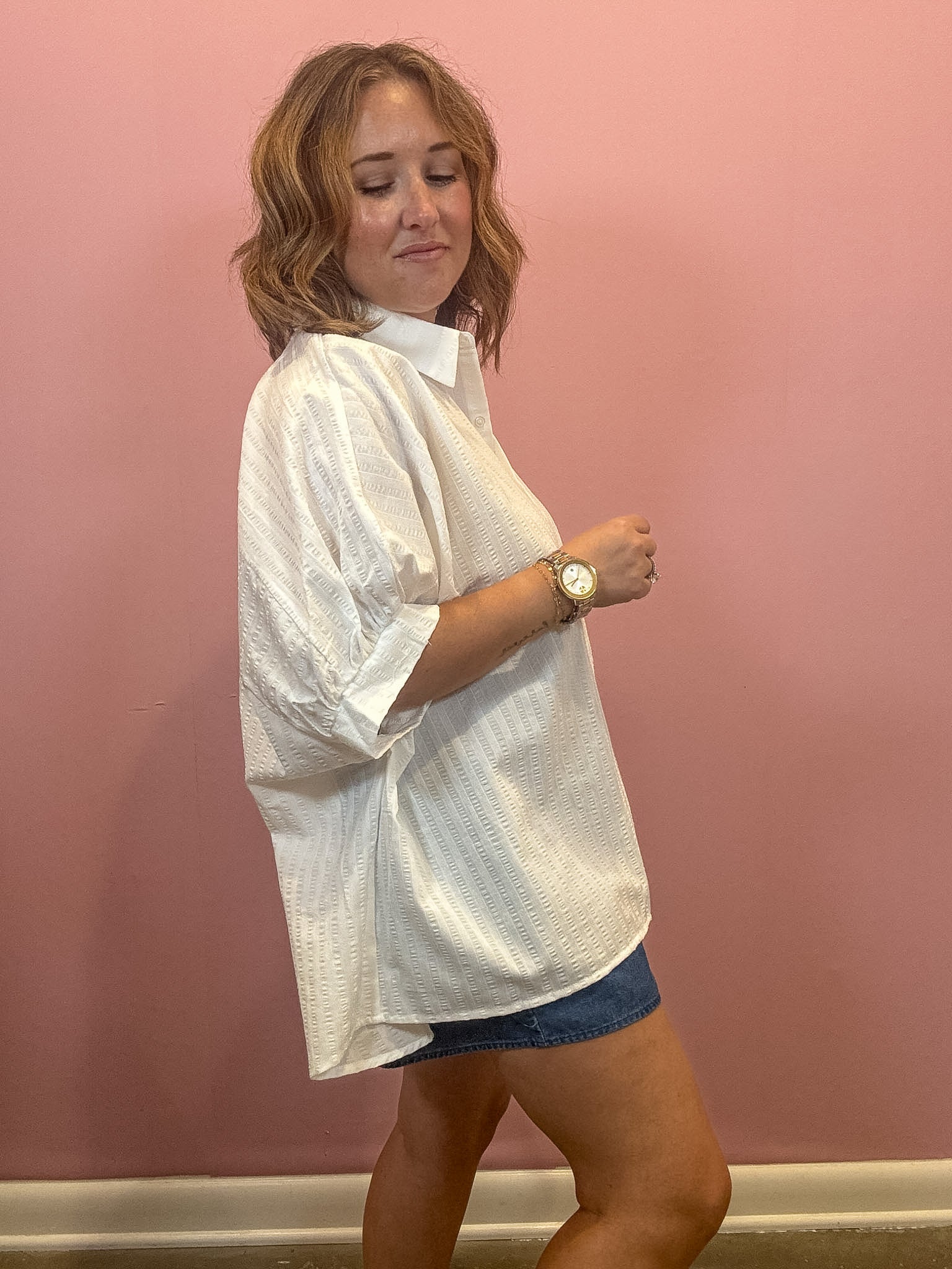A woman wearing an oversized white textured short-sleeve button-up shirt and denim shorts, standing against a solid pink wall.