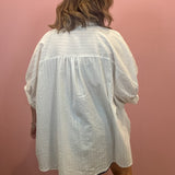 A person viewed from behind wearing an oversized white textured short-sleeve blouse or tunic in front of a pink wall.
