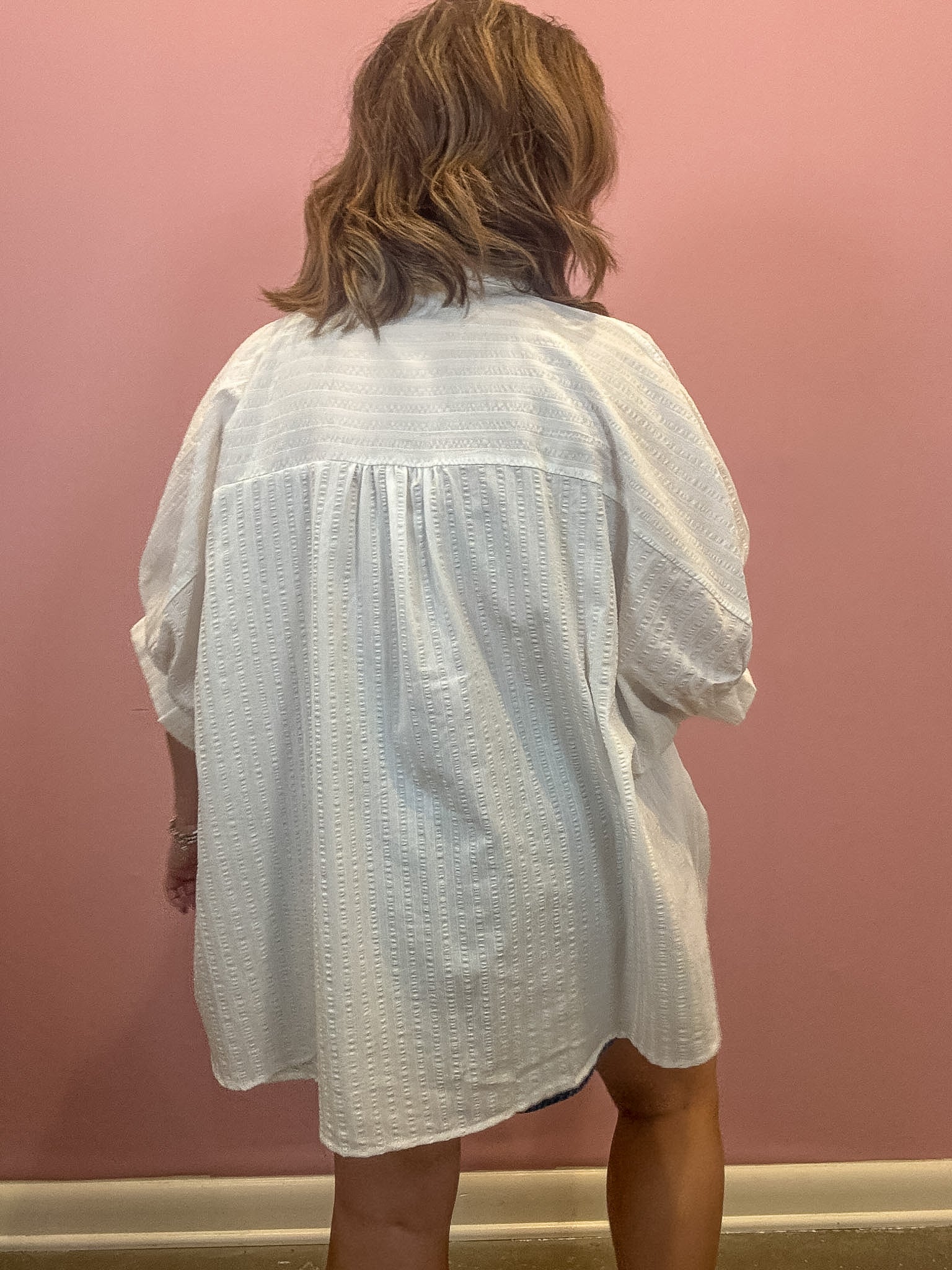 A person viewed from behind wearing an oversized white textured short-sleeve blouse or tunic in front of a pink wall.