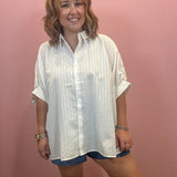A woman wearing a white textured short-sleeve button-up shirt and denim shorts, standing against a solid pink wall.