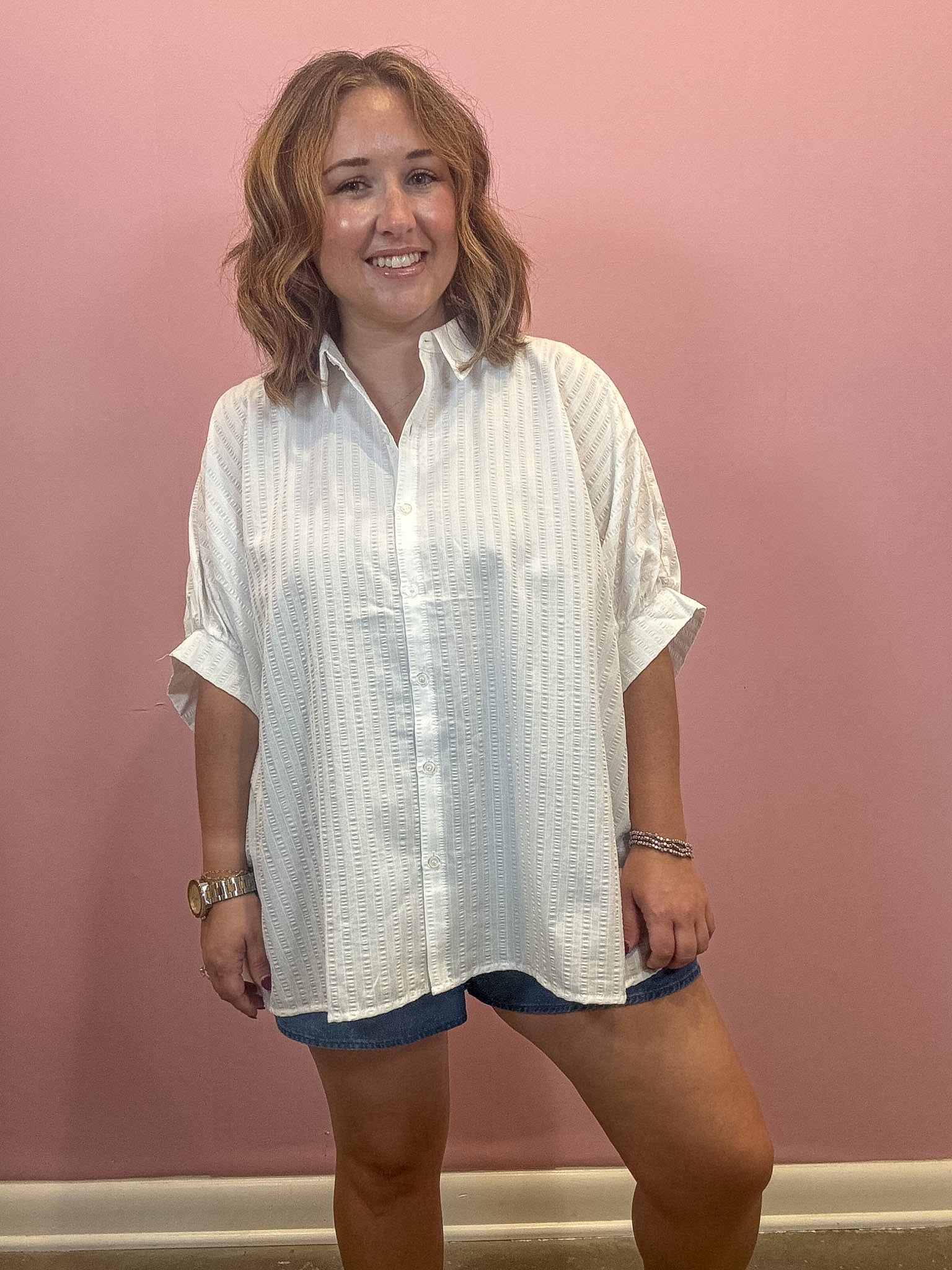 A woman wearing a white textured short-sleeve button-up shirt and denim shorts, standing against a solid pink wall.