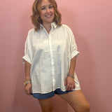 A woman modeling an oversized white textured button-down shirt with rolled sleeves, paired with denim shorts, standing against a solid pink wall.