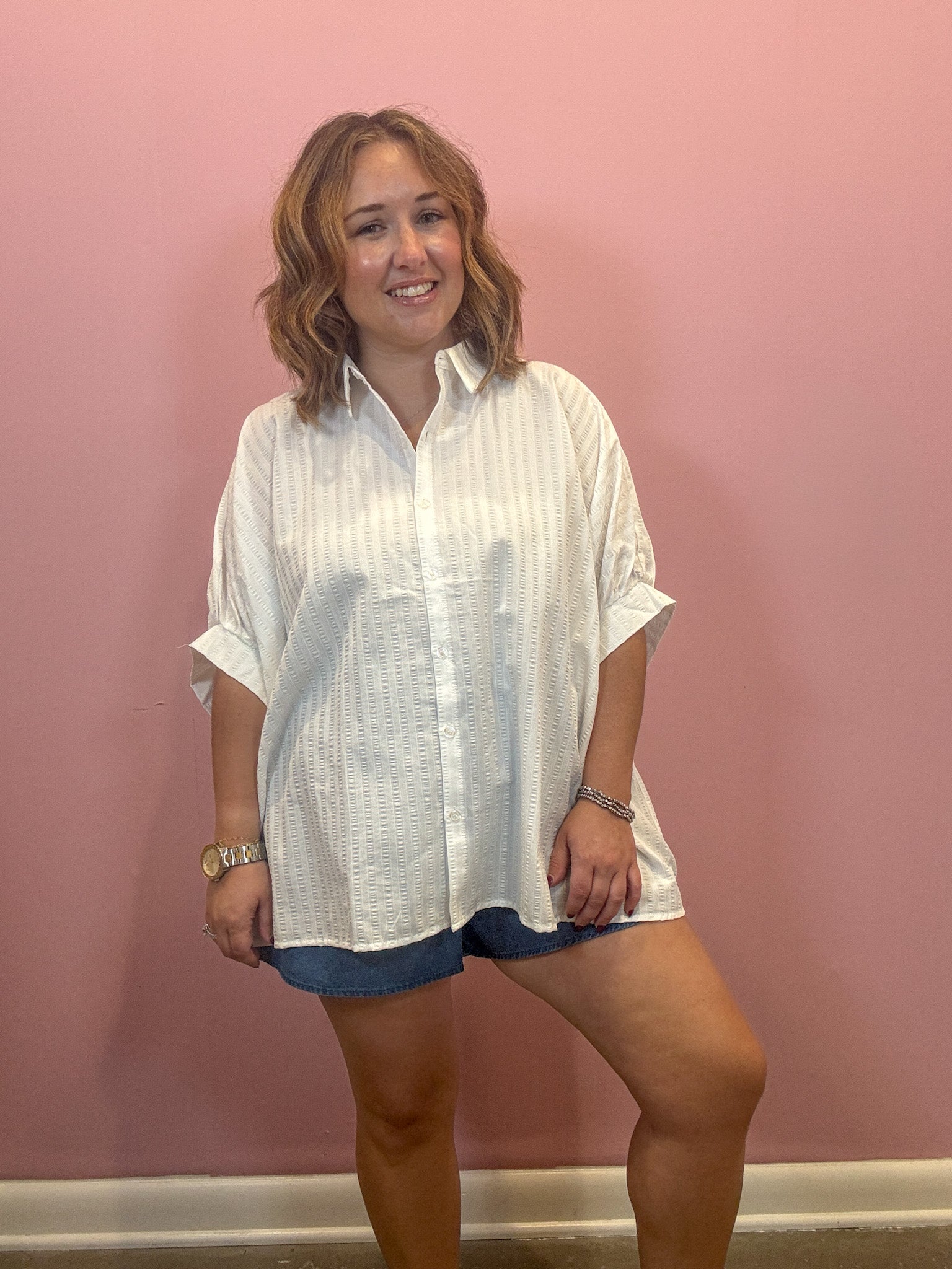 A woman modeling an oversized white textured button-down shirt with rolled sleeves, paired with denim shorts, standing against a solid pink wall.