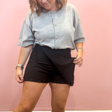 A woman wearing a light gray short-sleeve henley shirt and black shorts, standing against a solid pink wall.