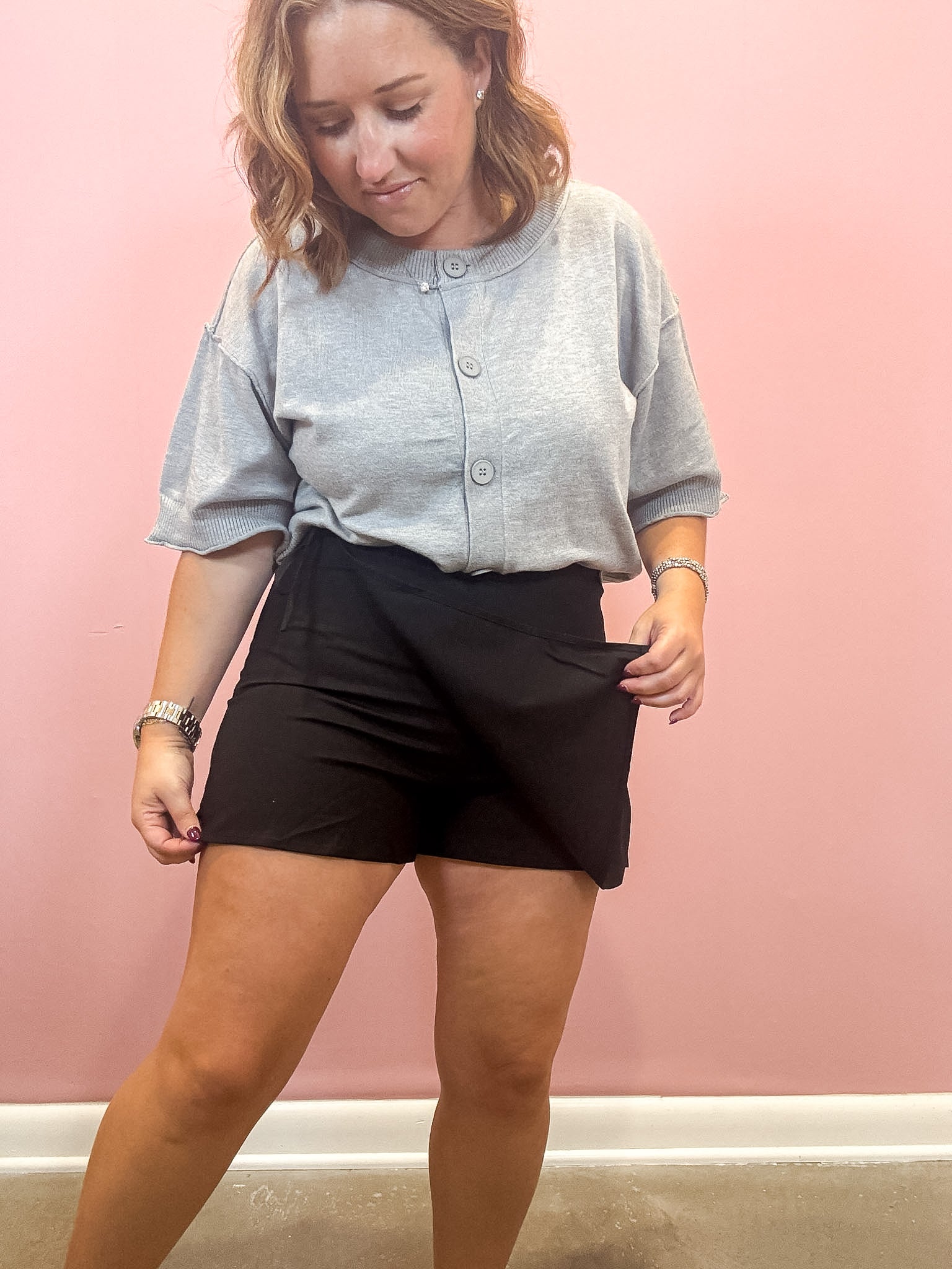 A woman wearing a light gray short-sleeve henley shirt and black shorts, standing against a solid pink wall.
