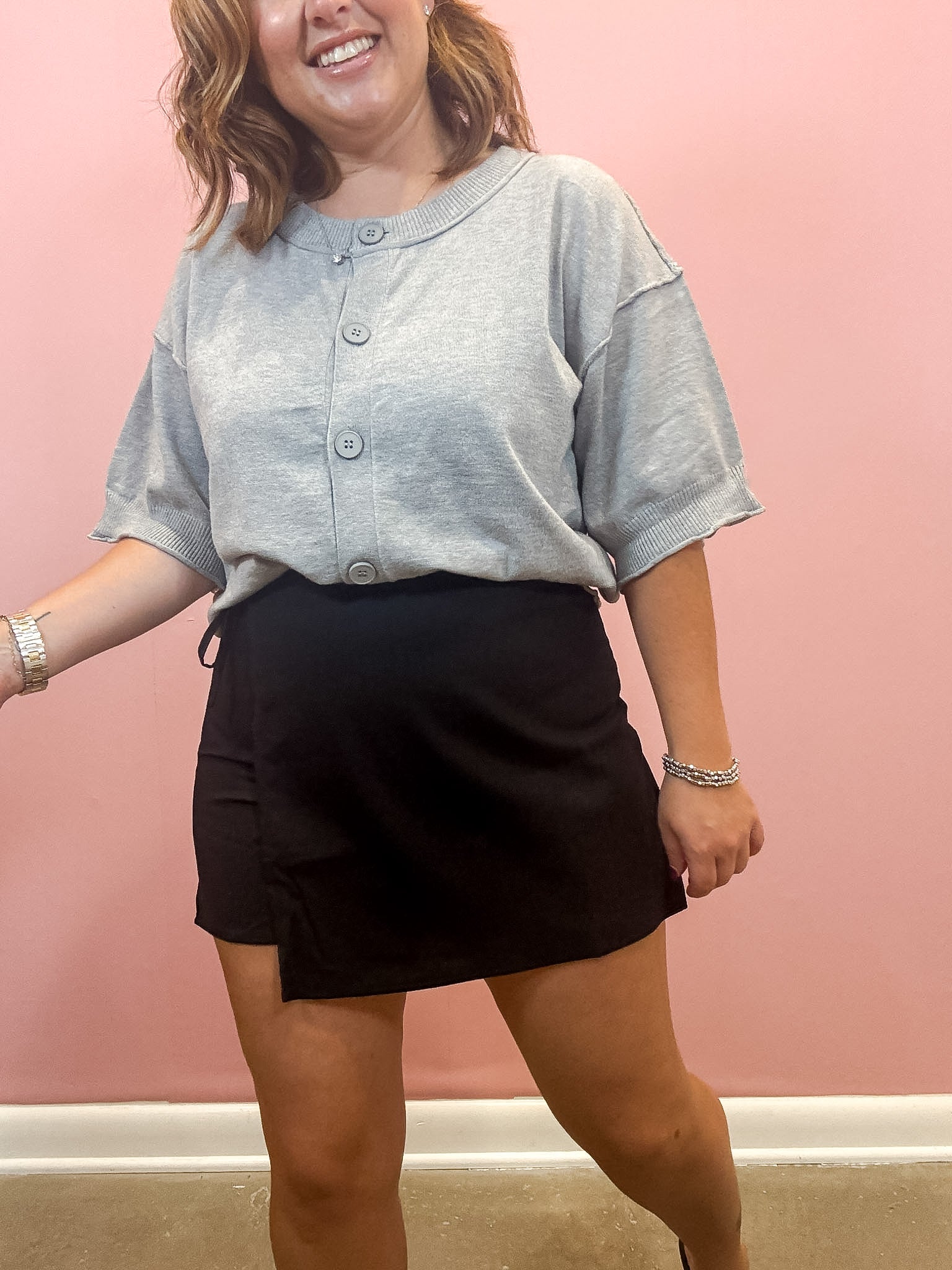 A woman wearing a light gray short-sleeve henley-style top and a black wrap-style mini skirt, standing against a solid pink wall.