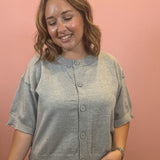 A woman wearing a light gray short-sleeve knit cardigan with a button-up front, standing against a solid pink wall.
