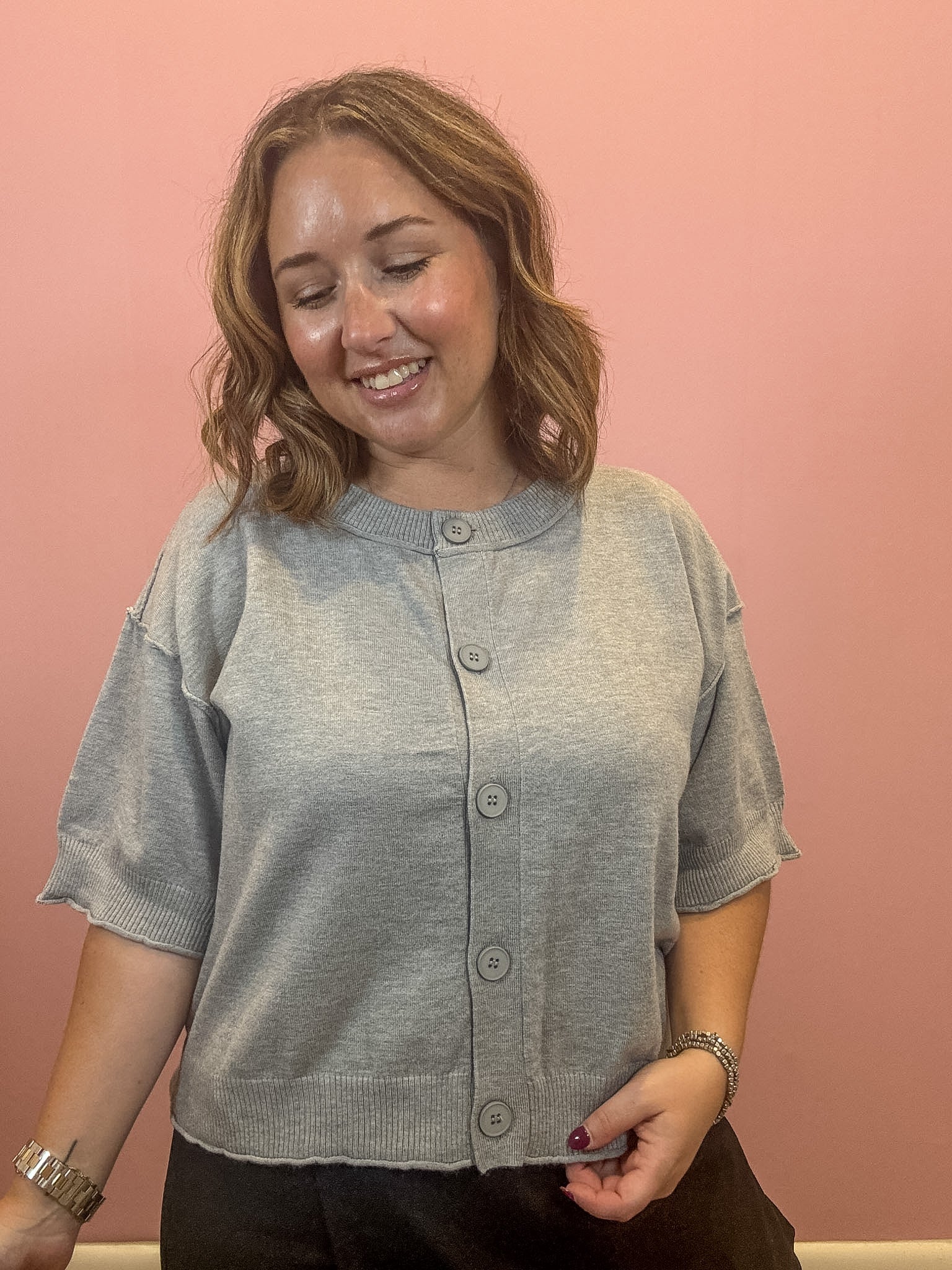 A woman wearing a light gray short-sleeve knit cardigan with a button-up front, standing against a solid pink wall.