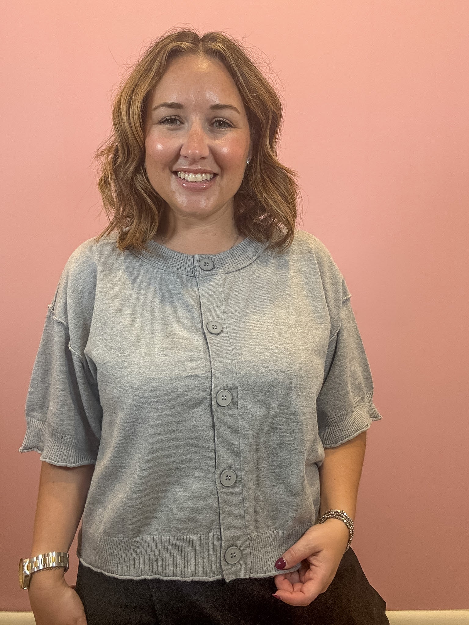 A smiling woman with shoulder-length brown hair wearing a light gray short-sleeve button-front cardigan, standing against a solid pink wall.