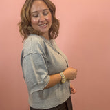 A woman with shoulder-length brown hair is shown from the side, wearing a light gray knit top and dark brown pants, standing against a solid pink wall.