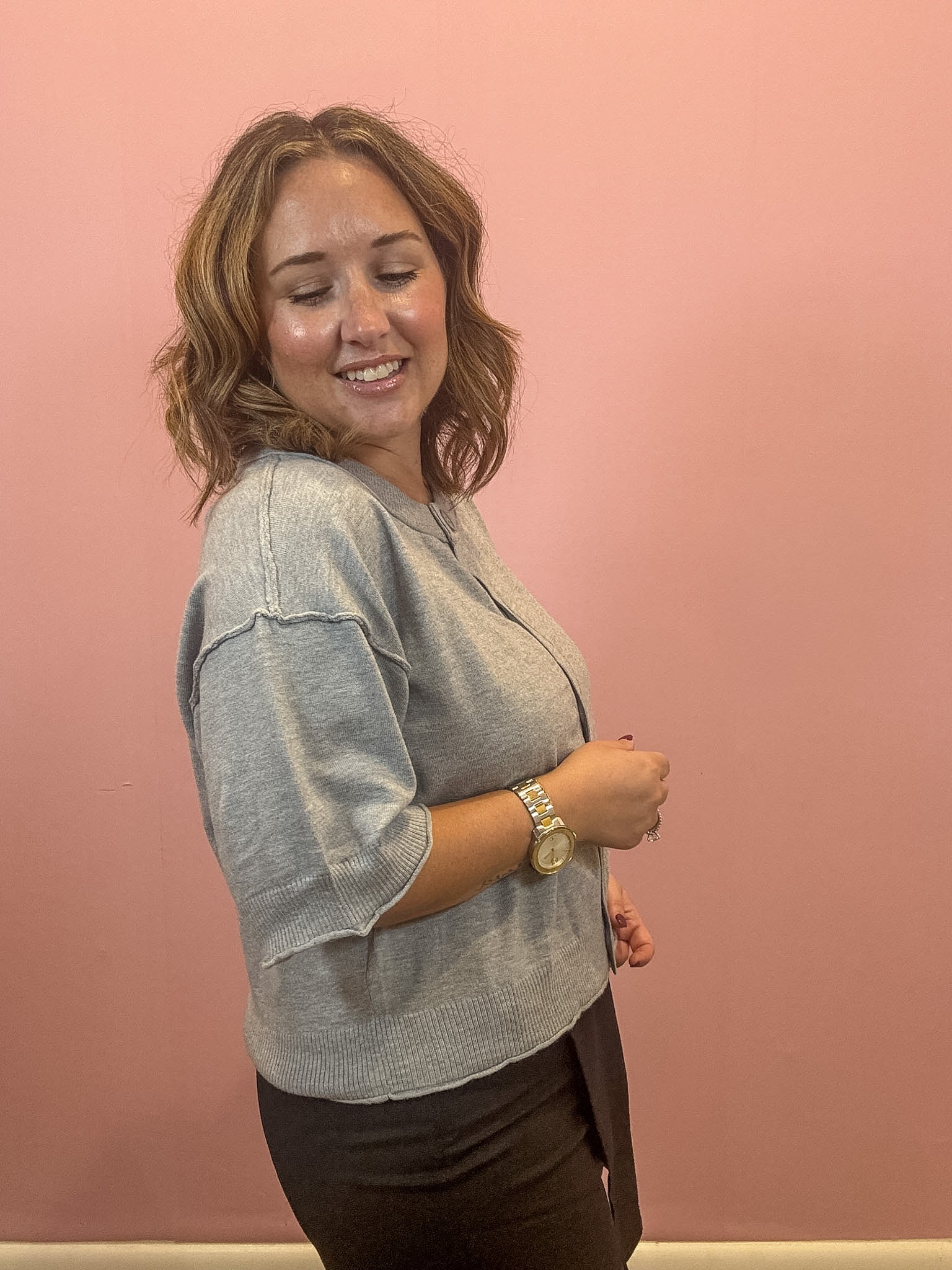 A woman with shoulder-length brown hair is shown from the side, wearing a light gray knit top and dark brown pants, standing against a solid pink wall.