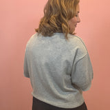 A woman with shoulder-length blonde hair is shown from the back wearing a light gray knit crewneck sweater and black shorts, standing against a solid pink wall.
