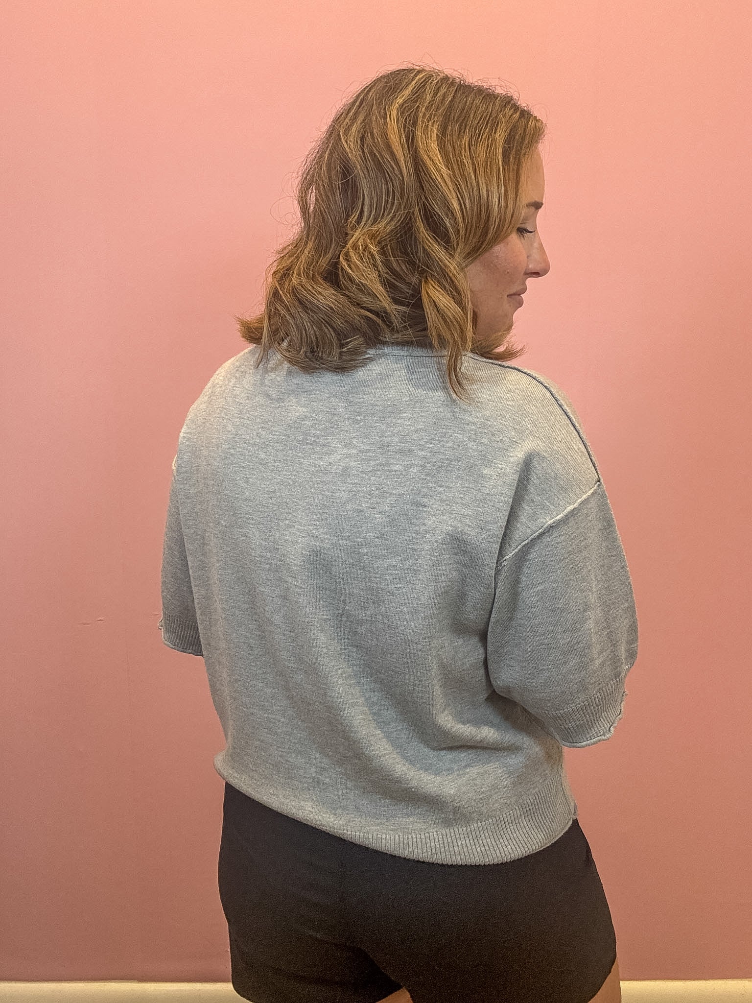 A woman with shoulder-length blonde hair is shown from the back wearing a light gray knit crewneck sweater and black shorts, standing against a solid pink wall.