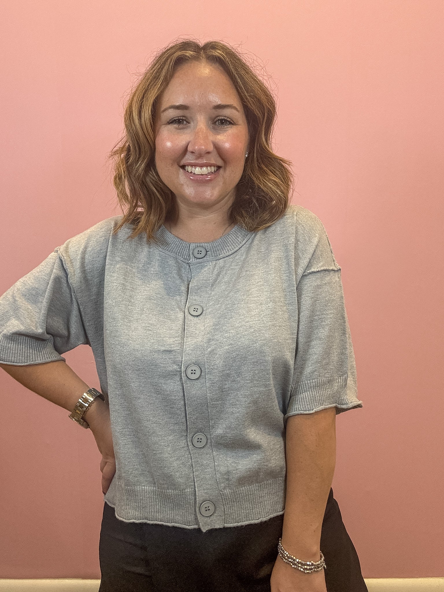A smiling woman with shoulder-length brown hair stands against a solid pink wall, wearing a light gray short-sleeve button-front cardigan and dark pants.