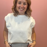 A smiling woman with shoulder-length blonde hair wearing a white sleeveless crewneck t-shirt and a black-and-white gingham patterned skirt, standing against a solid pink wall.